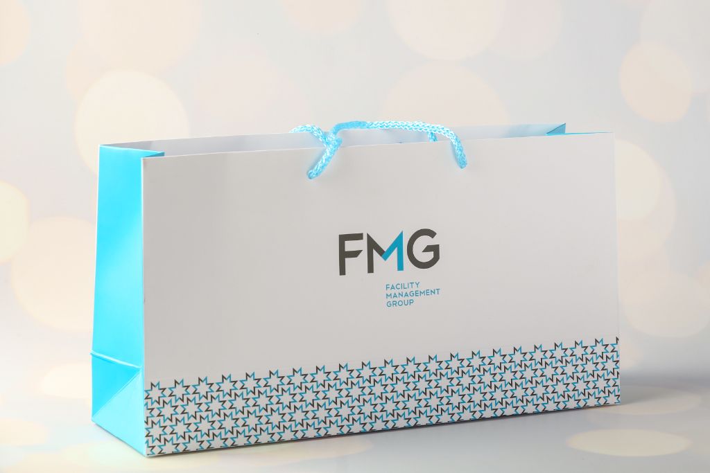 FMG Facility Management Group horizontal bag — white, teal sides, blue handle, ornamental border at bottom