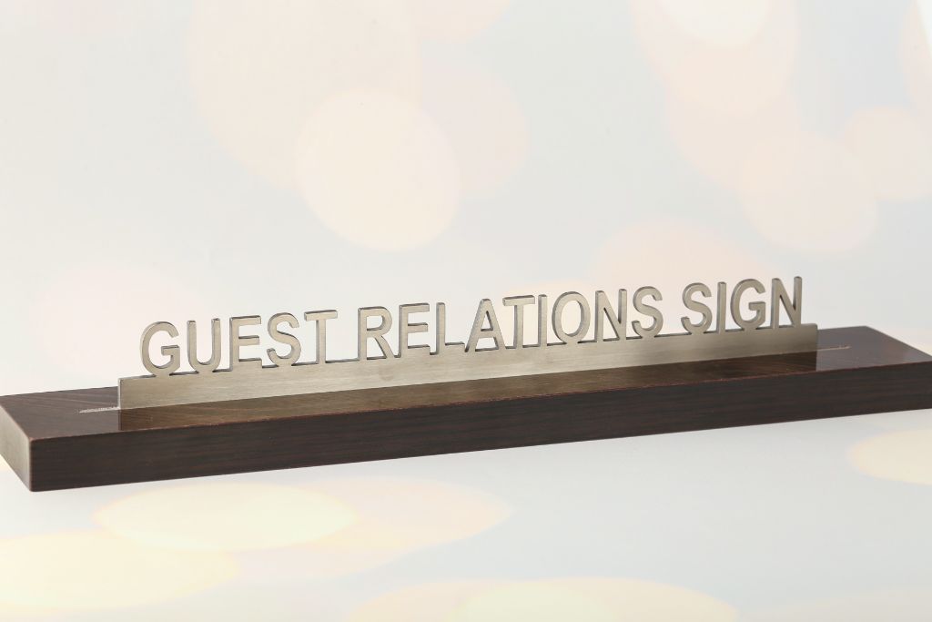 3D cut letters GUEST RELATIONS SIGN — front view, stainless steel, wenge wood base, reception desk area